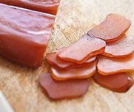 Sulla sinistra pezzo di bottarga di tonno, sulla destra fette di bottarga; il colore è quello tipico del tonno crudo, rosa quasi rosso; sotto piano in legno