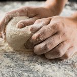 Male chef hands knead dough with flour on kitchen table