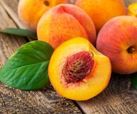 Su un tavolo in legno pesche intere dalla buccia aranciata screziata di rosso; in primo piano una metà di pesca a pasta gialla, con un nocciolo marrone