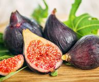 Alcuni fichi interi, una metà e un quarto; la buccia è di colore viola intenso e l’interno rosa-rosso; dietro, sfocate, alcune foglie, sotto un piano di legno