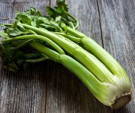 Un cespo di coste di sedano crudo con le sue foglie; la tipologia è verde, con steli color prato più chiari alla base; sotto, un piano in legno grigio