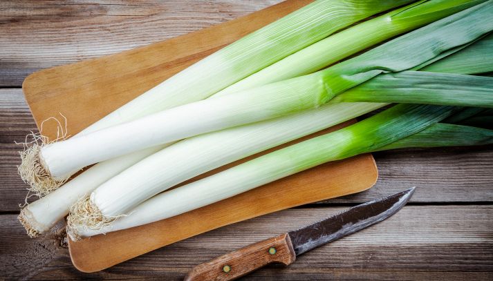 Su un tagliere rettangolare, cinque porri freschi con ancora la barbetta, pronti per essere puliti e cucinati; sul tavolo in legno c’è anche un coltello