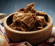 In una scodella di legno chiaro su uno strofinaccio a quadri gialli, neri e rossi, ciccioli rosolati in padella pronti per essere serviti. Sfondo grigio.