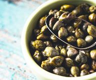 In primo piano, sulla destra, una scodella di ceramica bianca e un cucchiaio in metallo, entrambi pieni di capperi freschi; sfondo azzurrino sulla sinistra