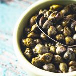 In primo piano, sulla destra, una scodella di ceramica bianca e un cucchiaio in metallo, entrambi pieni di capperi freschi; sfondo azzurrino sulla sinistra