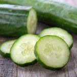 Davanti, in centro, alcune fette di cetriolo dalla polpa quasi bianca e con la buccia liscia verde scuro; dietro un cetriolo tagliato a metà e uno intero