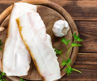 Da sinistra a destra, vista dall’alto di due fette di lardo dalla consistenza umida e soffice, testa d’aglio, prezzemolo; il tutto su tagliere rotondo su tavolo in legno