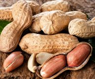 Arachidi da guscio color beige, con semi oblunghi ricoperti da una pellicina sottile rosa-rossa; sotto tavolo in legno