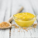 Una ciotola rotonda di vetro riempita con salsa di senape e un cucchiaio di legno con semi, entrambi appoggiati su un tavolo di legno bianco