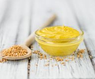 Una ciotola rotonda di vetro riempita con salsa di senape e un cucchiaio di legno con semi, entrambi appoggiati su un tavolo di legno bianco