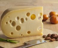 Un pezzo di Emmental dal color giallo chiaro con grandi buchi nella pasta su tagliere di legno; in basso erba cipollina, coltello e noci pecan