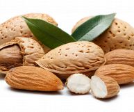 Mandorle con guscio, mandorle con buccia color cammello e una mandorla spezzata a metà, dalla polpa color avorio; sopra due foglie lucide verdi scuro