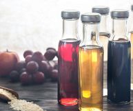 Cinque bottiglie trasparenti con aceti di vario genere; sulla sinistra uva rossa, prugne e un cucchiaio in legno con riso crudo