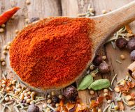 In primo piano, paprika dolce su cucchiaio di legno; sul tavolo peperoncino, stecca di cannella, cardamomo, semi di finocchio, senape, ginepro