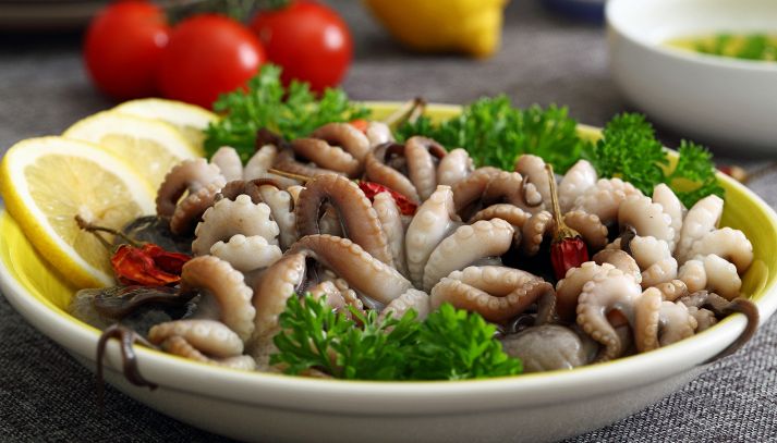 Moscardini in insalata, con tentacoli rosati sulla punte; nello stesso piatto, foglie intere di prezzemolo e spicchi di limone; sfondo sfocato con pomodori Piccadilly