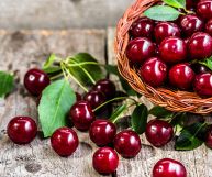 Sulla destra cestino di vimini con ciliegie rosso vivo, sulla sinistra amarene sparse con foglie verdi; il tutto appoggiato su un tavolo in legno