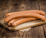 Quattro wurstel lunghi e sottili, probabilmente tedeschi. Sono pronti per essere cucinati e sono posati su un tagliere di legno chiaro dalla forma rettangolare