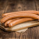 Quattro wurstel lunghi e sottili, probabilmente tedeschi. Sono pronti per essere cucinati e sono posati su un tagliere di legno chiaro dalla forma rettangolare