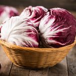 In un cestino di vimini marrone chiaro tre teste di radicchio di Chioggia; dietro, sfocate e su piano in legno, altre teste di radicchio bianche e viola
