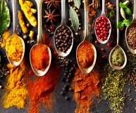 Cucchiaini affiancati su sfondo nero, riempiti con coriandolo, zenzero, paprika, peperoncino, pepe nero, pepe rosa, semi di cumino e di finocchio. Spezie sparse