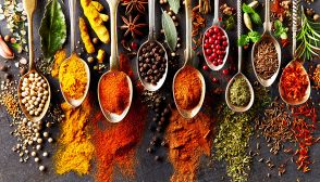 Cucchiaini affiancati su sfondo nero, riempiti con coriandolo, zenzero, paprika, peperoncino, pepe nero, pepe rosa, semi di cumino e di finocchio. Spezie sparse