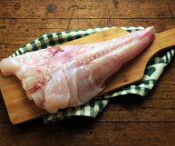 Su un piccolo tagliere, un filetto di coda di rospo senza pelle né lische; sotto uno panno a quadri bianchi e verdi poggiato su un tavolo in legno