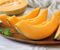 Su un piatto rotondo di legno, fette di melone con scorza increspata, polpa arancione e semini beige; dietro a sinistra la restante metà del melone