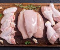 Da sx a dx, quattro ali di pollo, tre petti e quattro cosce di pollo pronte per essere cucinate; erbe aromatiche e aglio; il tutto su un tagliere