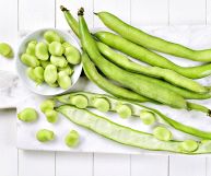 Sulla destra alcuni gusci di fave fresche per la maggior parte chiusi, sulla sinistra una scodella di fave sgranate; dietro un tagliere bianco su un tavolo chiaro