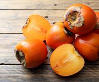 Su un tavolo in legno sono appoggiati quattro cachimela interi e uno aperto in due metà; il colore è arancio brillante e la pelle liscia e tesa