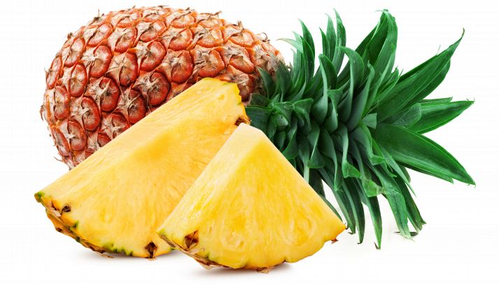 Su sfondo bianco, due fette di ananas mature con scorza; dietro un frutto intero dalla buccia aranciata, con foglie verdi brillante