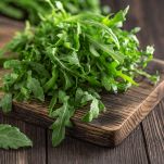 Una manciata di foglie di rucola verde intenso, lavate e pronte all'uso, posate su un tagliere di legno su piano dello stesso materiale