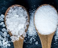 Su sfondo scuro in ardesia la punta di due cucchiai di legno chiaro; quello di sinistra è riempito con sale grosso mentre quelle di destra con sale fino