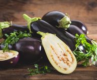 Su un tavolo e uno sfondo marrone, mucchio di melanzane allungate dalla buccia lucida viola scuro; è presente il picciolo verde e una è tagliata a metà