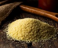 Una montagnola di couscous da cuocere su un tavolo di legno in primo piano; dietro, sulla sinistra della iuta, sulla destra un cucchiaio in legno