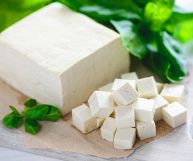 Una forma rettangolare di tofu in parte tagliata a cubetti, dalla pasta soda di colore bianco; è adagiato su carta oleata beige e un tavolo di legno chiaro