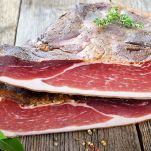 Due metà di speck su un piano rustico di legno; l'interno è rosso con parti più scure e porzioni equilibrate di grasso; l'esterno e bruno, senape e marrone