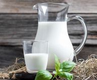 Latte intero in un bicchiere e in una caraffa di vetro appoggiati su paglia su cui si trova anche un ciuffo di foglie di basilico. Sfondo in legno