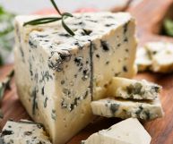Un pezzo di gorgonzola con pasta soda dal color avorio e la caratteristica erborinatura blu; sotto un tagliere di legno con altri pezzetti di formaggio