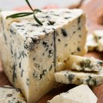 Un pezzo di gorgonzola con pasta soda dal color avorio e la caratteristica erborinatura blu; sotto un tagliere di legno con altri pezzetti di formaggio
