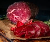 SU un tagliere in primo piano, fette di bresaola affumicata; sullo sfondo un pezzo tagliato a metà; ambientazione rustica per un tipico antipasto italiano