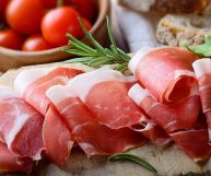 Fette di prosciutto crudo adagiate le une sulle altre e non schiacciate, su tagliere in legno con rosmarino; dietro pomodori ciliegini rosso brillante