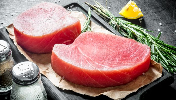 Due tranci di tonno rosso spessi 2 cm, pronti per essere cotti e insaporiti con sale, limone e rosmarino, condimenti posati sul tavolo nero