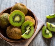 Kiwi interi con buccia marrone e kiwi tagliati a metà o in quarti con polpa verde brillante in una ciotola di legno sulla sinistra; altri pezzi di kiwi sulla destra; lo sfondo è gigio cenere