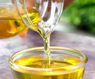 Una caraffa in vetro con olio di oliva che cade in una scodella dalle piccole dimensioni, anch’essa in vetro. Piano in pietra ed erbe aromatiche sullo sfondo