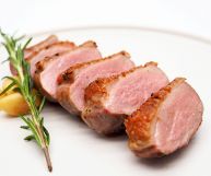 Un petto d’anatra tagliato in fette spesse, dalle carni succose dall’interno rosato e l’esterno rosolato;sotto piatto bianco, di fianco rosmarino