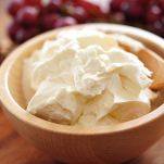 Mascarpone cremoso in una ciotola di legno chiaro, con un cucchiaio dello stesso materiale; sullo sfondo, sfocato, un grappolo di uva nera