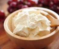 Mascarpone cremoso in una ciotola di legno chiaro, con un cucchiaio dello stesso materiale; sullo sfondo, sfocato, un grappolo di uva nera