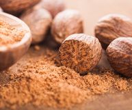 Noci moscate intere e macinate; la polvere è contenuto in una scodella di legno sulla sx, i pezzi integri sono adagiati su un fondo marrone sulla dx