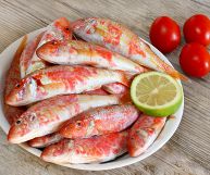 Un piatto di triglie fresche con una metà di un limone, su un piano in legno con pomodori; I pesci sono interi e freschi, ancora da cuocere